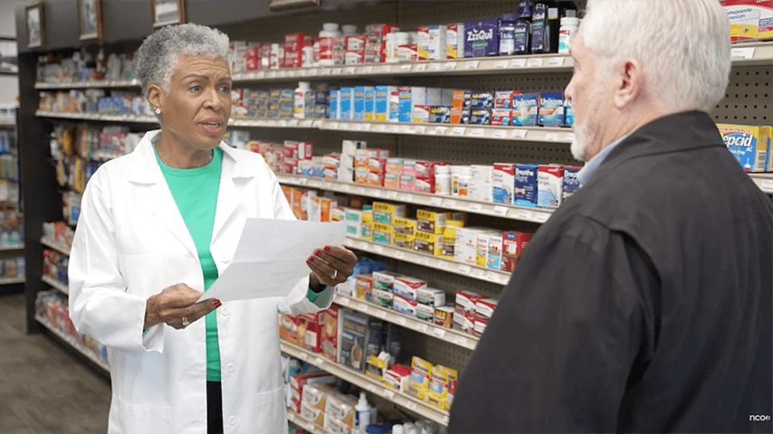 woman in lab coat talking to older man in pharmacy aisle