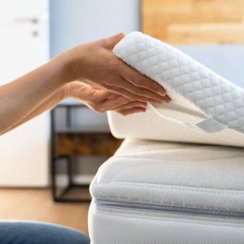 A woman lifts a mattress topper off a mattress slightly at the corner