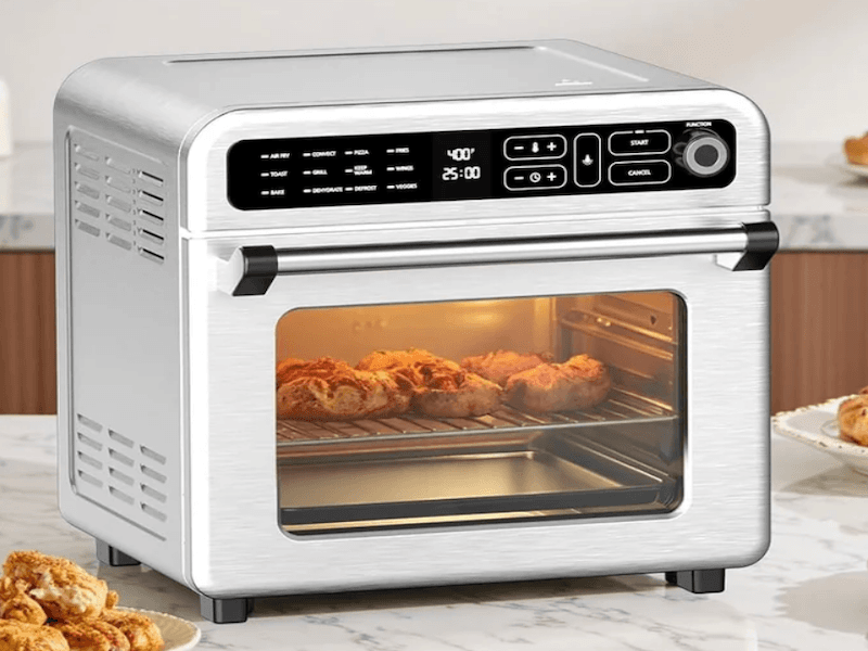 Modern silver air fryer oven on a kitchen countertop, glass front revealing pastries baking inside. The digital display shows time and temperature