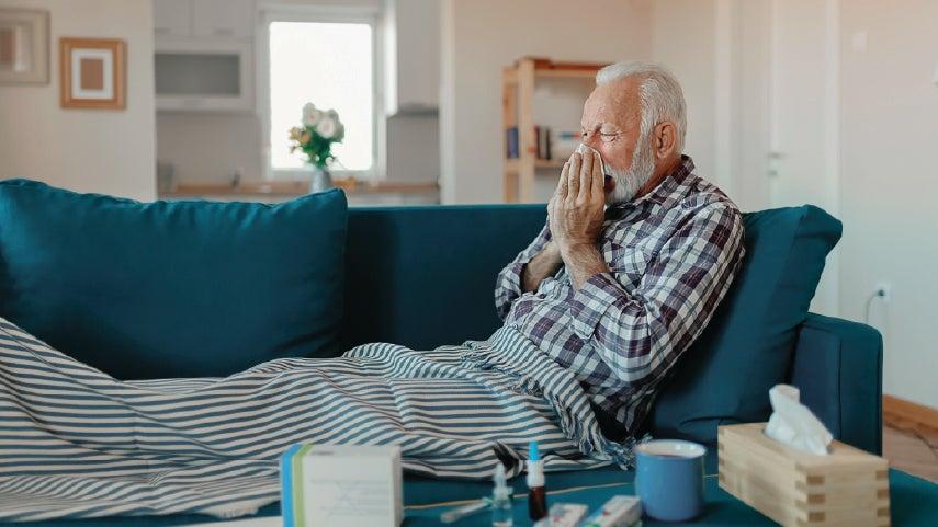 White senior man is sitting on the couch blowing his nose. He may have a cold or RSV.