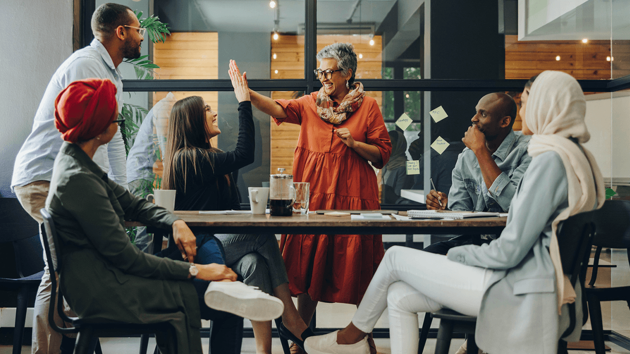 A diverse group of people is gathered around a table in a modern office setting. The group includes men and women of various ages and ethnicities. One woman with short gray hair and glasses, dressed in an orange dress and a colorful scarf, is standing and giving a high-five to a seated woman with long brown hair and glasses, who is smiling. The others are sitting around the table, engaged in the discussion. The background features glass walls with sticky notes, and the atmosphere appears collaborative and positive.