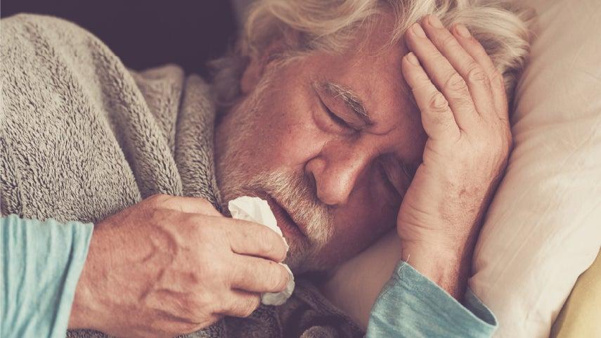 A senior man is laying in bed under his covers with a tissue close to his mouth and his eyes closed, suffering from covid.