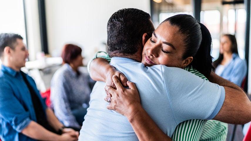 A Hispanic younger woman is giving an older senior man a hug in a group therapy session.
