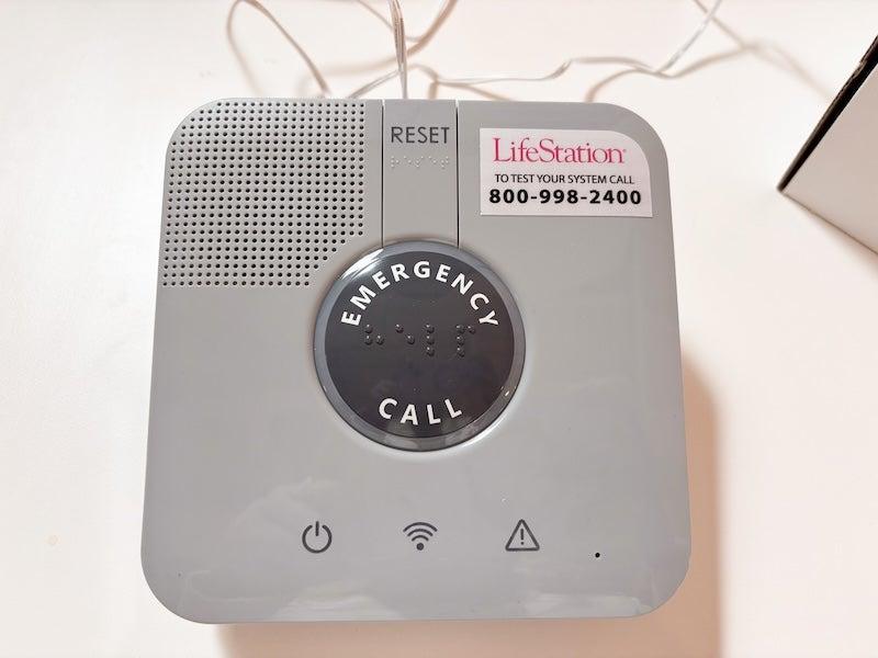 The LifeStation Sidekick Home base is a grey square with rounded corners. There is a circular black emergency call button at the center of the square, and tiny holes for a speaker in the upper-left corner.