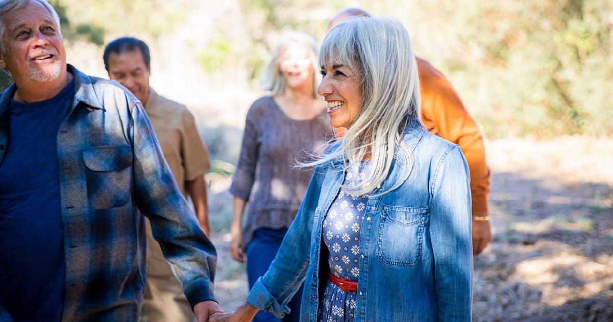 A group of older adults enjoying an outdoor walk together, smiling and holding hands. The focus is on a woman with long gray hair wearing a denim jacket and a floral dress, looking content and happy.