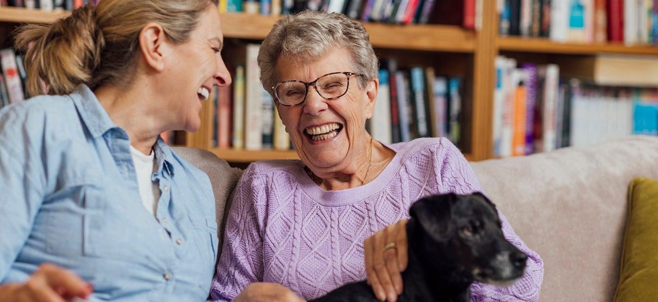 Two women sitting on a couch and laughing with a dog on one of their laps.