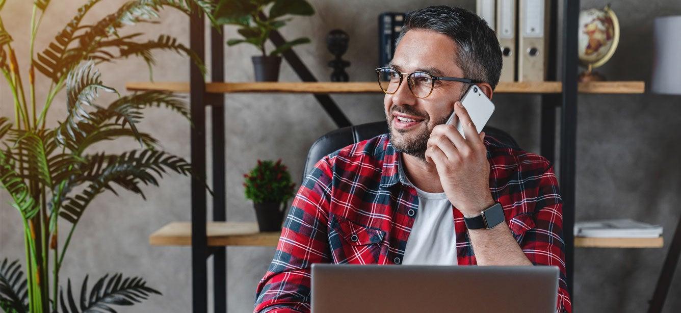 Smiling man in a plaid shirt sitting at a desk with a laptop, talking on the phone in a modern office with plants.