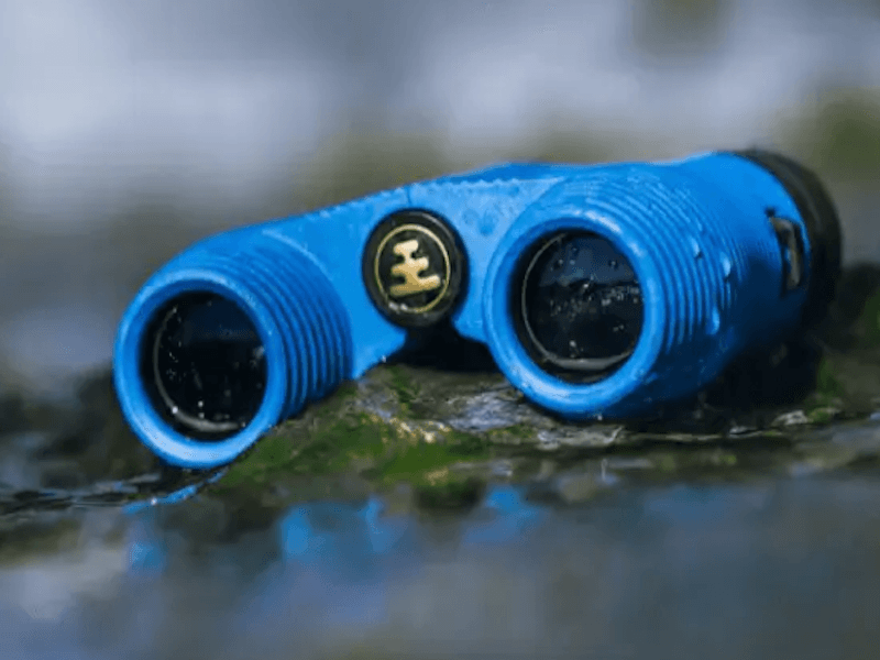 Blue pair of binoculars on natural surface, covered with beads of moisture to show they’re waterproof
