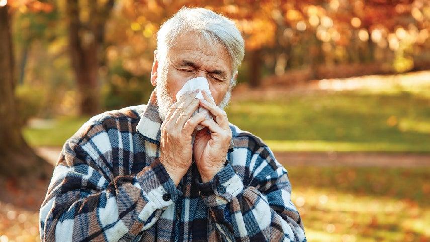Older gentleman blowing his nose outside, with his eyes closed.