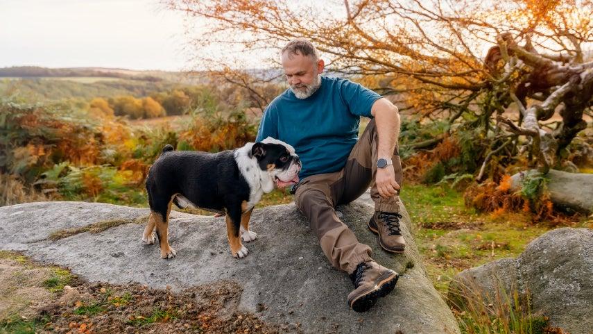A senior caucasian man is sitting outside on a mountain, overlooking the view with his bulldog.
