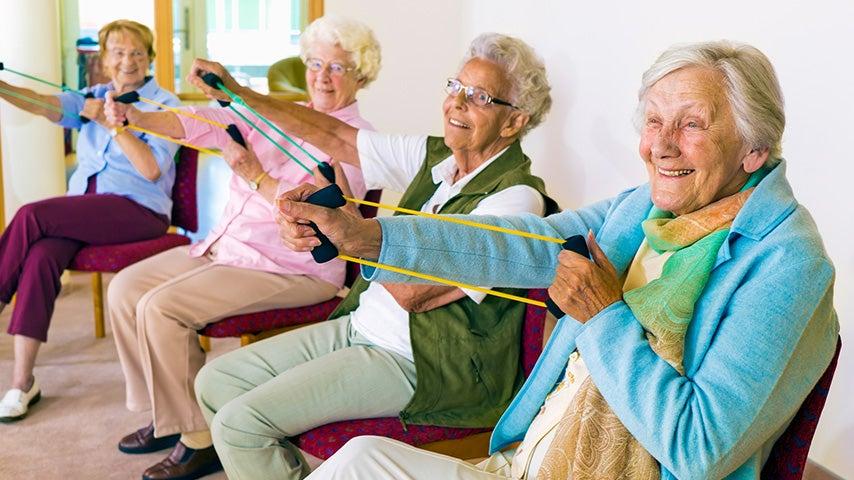 A group of women sit in chairs and use exercise bands to stretch their arms.