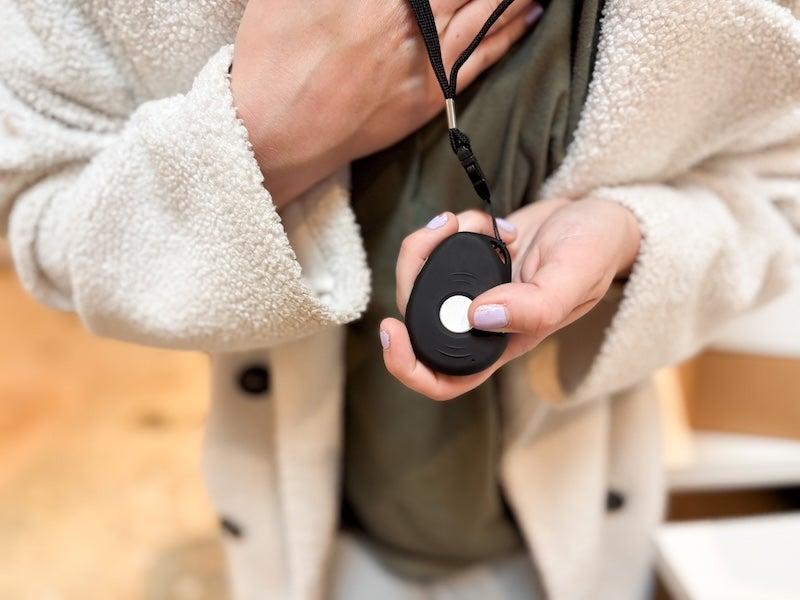 A tester displays a close-up of the chunky black LifeStation Sidekick necklace, roughly the size of a fob for car keys, on its black lanyard attachment. The tester wears a white sherpa jacket and lavender nail polish on short nails.