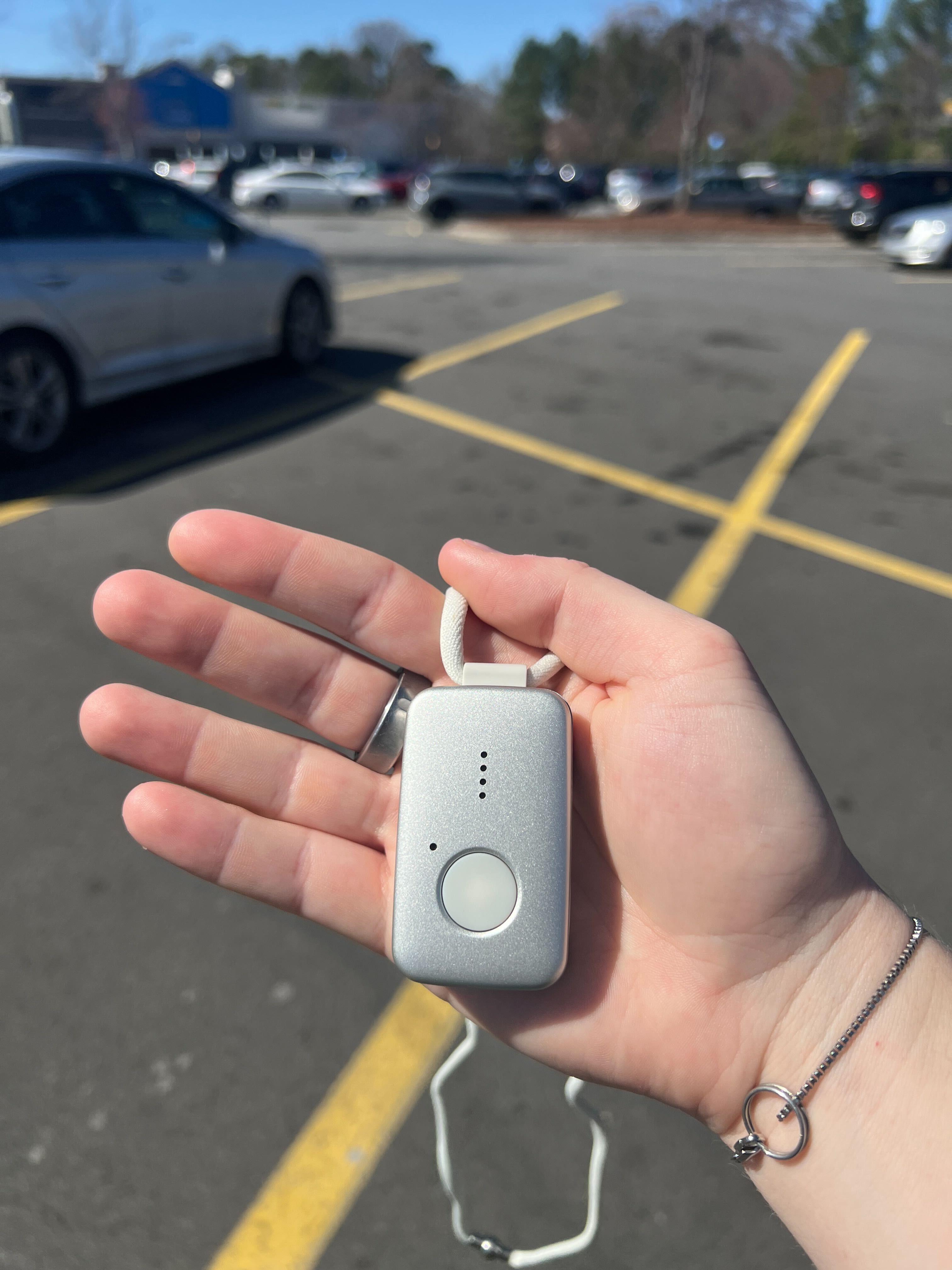 A person holds a small grey medical alert help button with a parking lot in the background