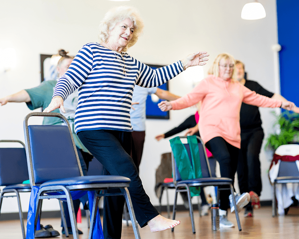 Older adults participate in a balance exercise class, using chairs for support during a standing exercise.