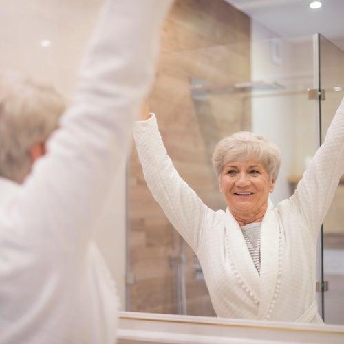 A happy woman stands in front of a bathroom mirror with her arms raised