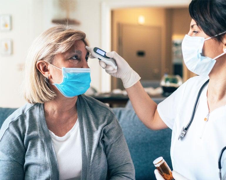 A masked doctor is taking the temperature of a senior masked woman in her home during the pandemic.