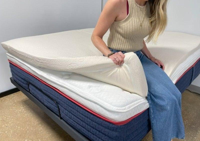 A woman sitting on the side of the Avocado mattress topper, lifting its corner