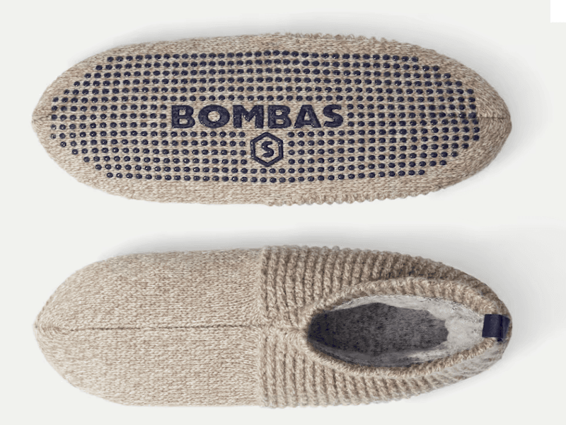 Tan-colored slippers with a wool interior and a grippy bottom, showing the “Bombas” logo on the bottom of one slipper