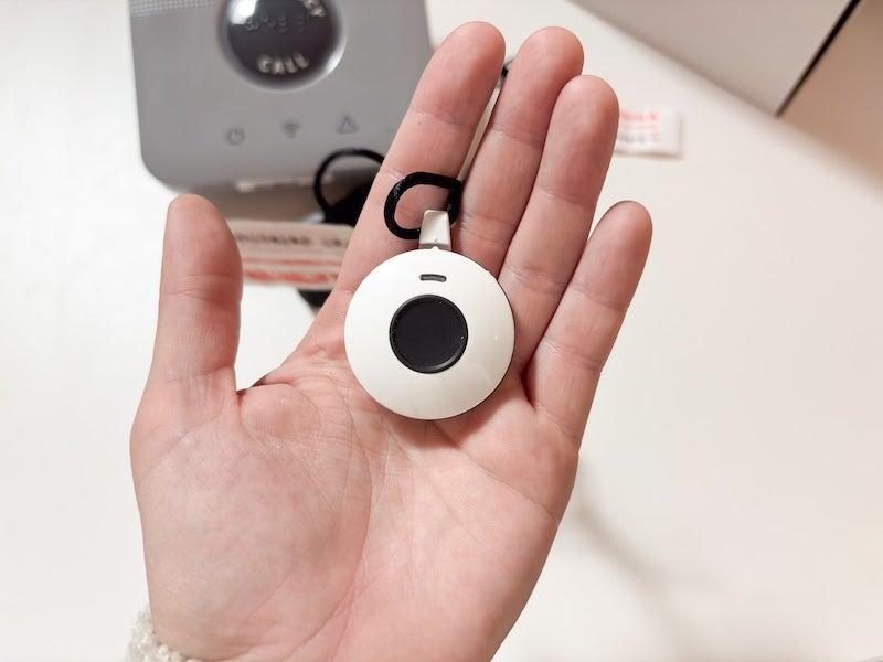 A person holds the white, circular LifeStation necklace in their open left palm. The necklace is the size of a half-dollar, and has a black necklace cord and a dime-sized black button in the center.