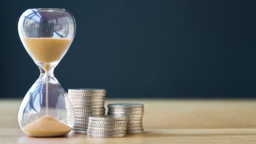 A close up of an hour glass pouring sand, indicating that time is passing - with coins next to it.