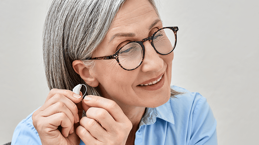 smiling woman with gray hair and glasses putting hearing aid into left ear