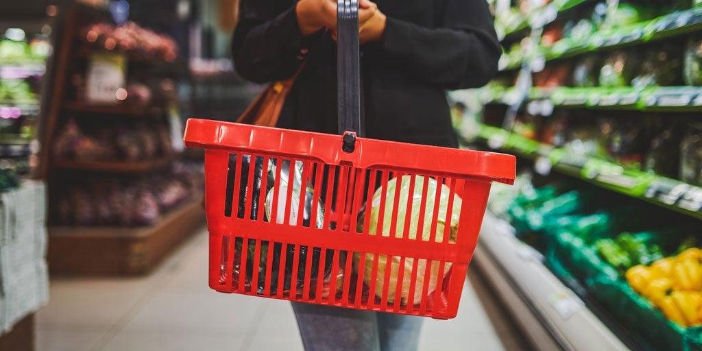 A female is holding a red grocery basket with produce in it in a aisle at the grocery store.