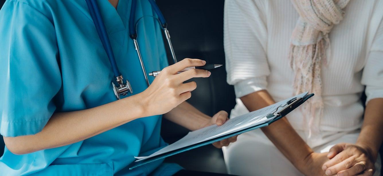 Healthcare provider in scrubs holds a clipboard and a pen while sitting with a patient.