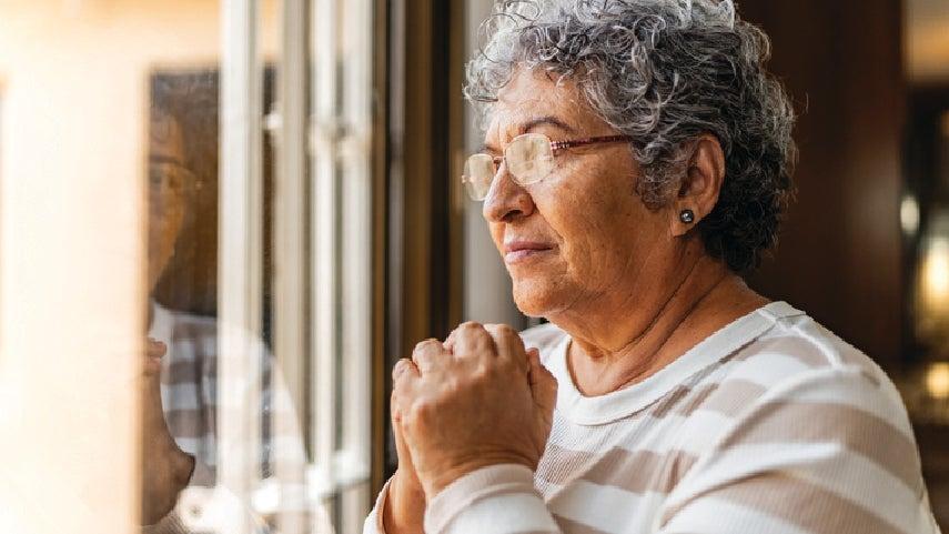 A woman looks out of a window with her hands clasped in front of her.
