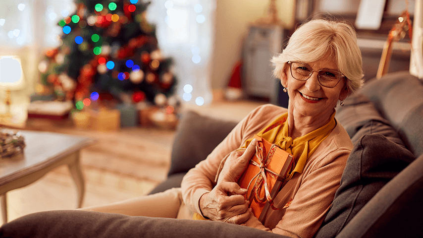 smiling older woman holding present with lighted Christmas tree in background