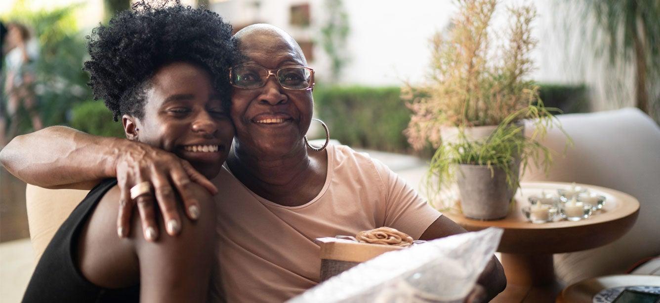 A young Black female caregiver is giving her grandmother a big hug in her home.