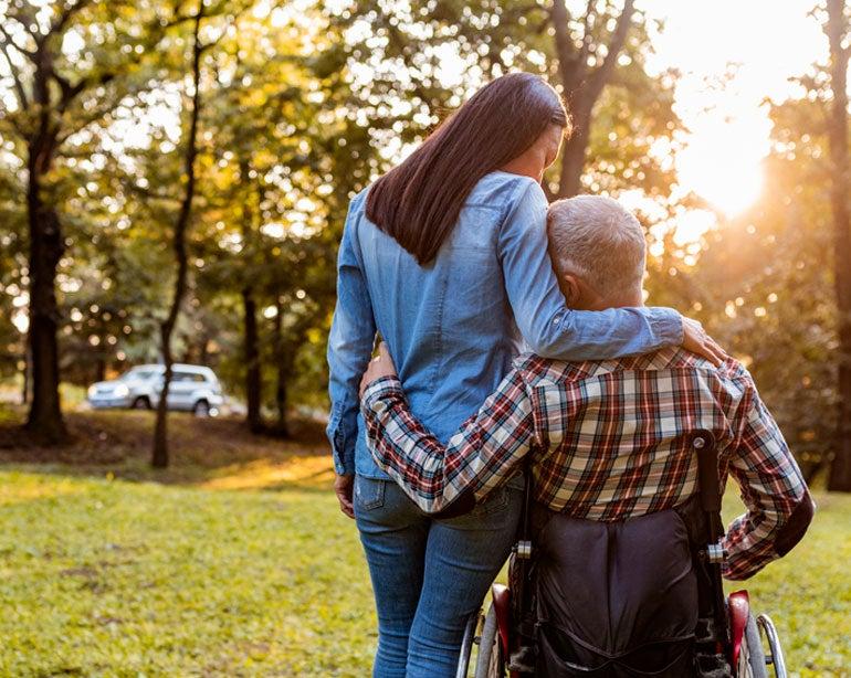A senior man in a wheelchair is hugging his younger caregiver while they're outside at a park.