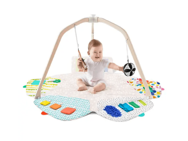 Baby sitting on a play mat and reaching for toys hanging from a wooden frame.