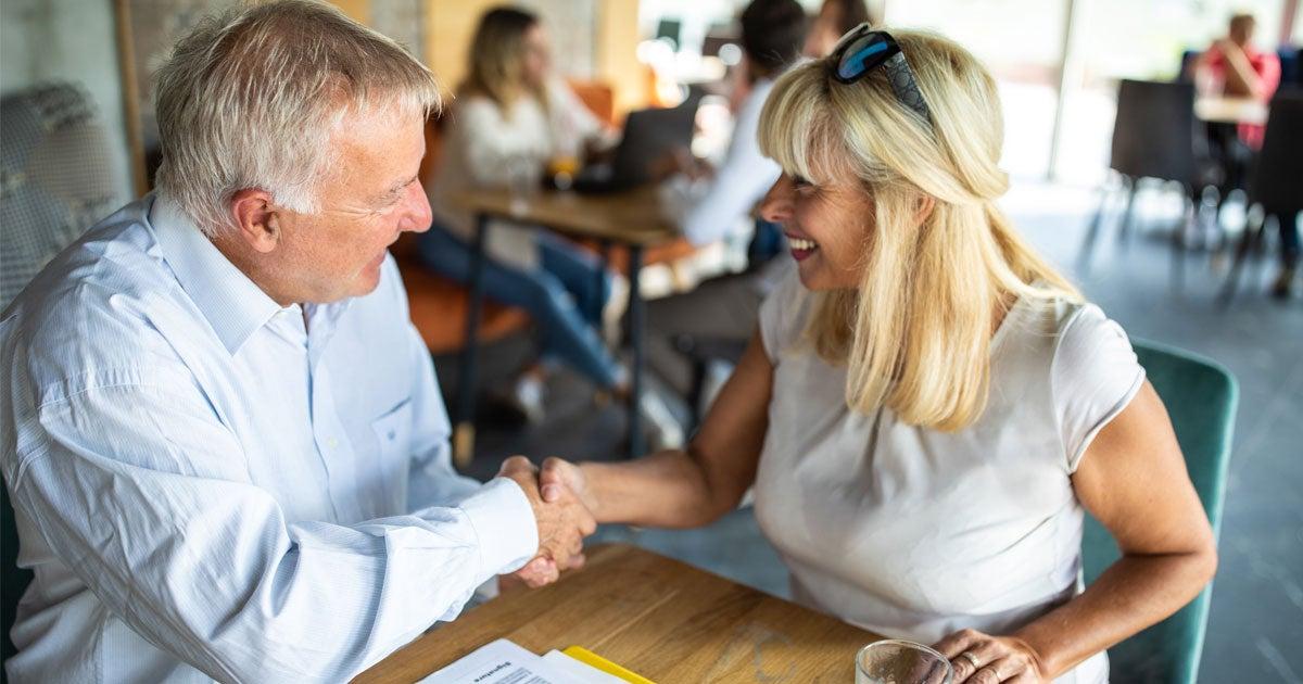 A senior Caucasian female shakes hands with a senior Caucasian man. Both are sitting at a table, discussing a financial agreement.