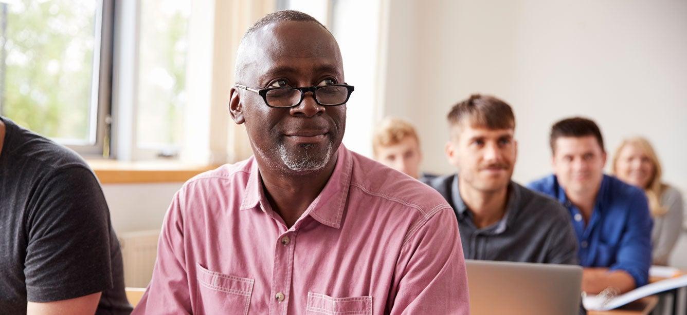 Man in glasses and a pink shirt sitting in a classroom with a group of adults listening attentively in the background.