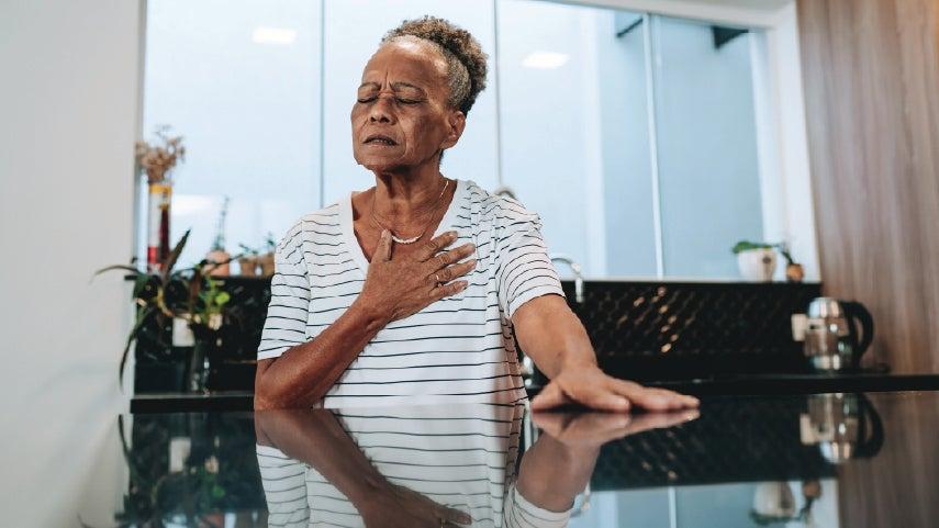 An older Black woman is sitting at her kitchen table clutching her chest with her hand with her eyes closed.