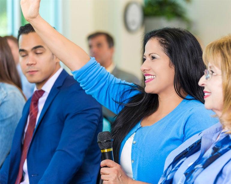 A caregiver to an older adult holds a mic at a town hall meeting as she prepares to ask a question.