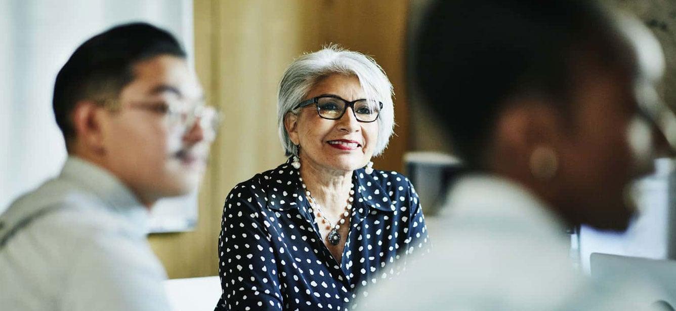 A senior female with glasses on sits in a room of professionals during a meeting.