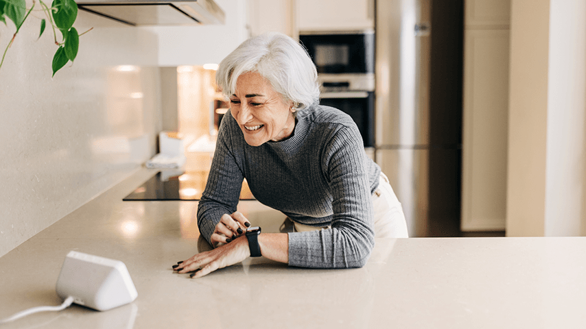 smiling woman with gray hair leaning on kitchen counter looking at smart home device while adjusting smart watch