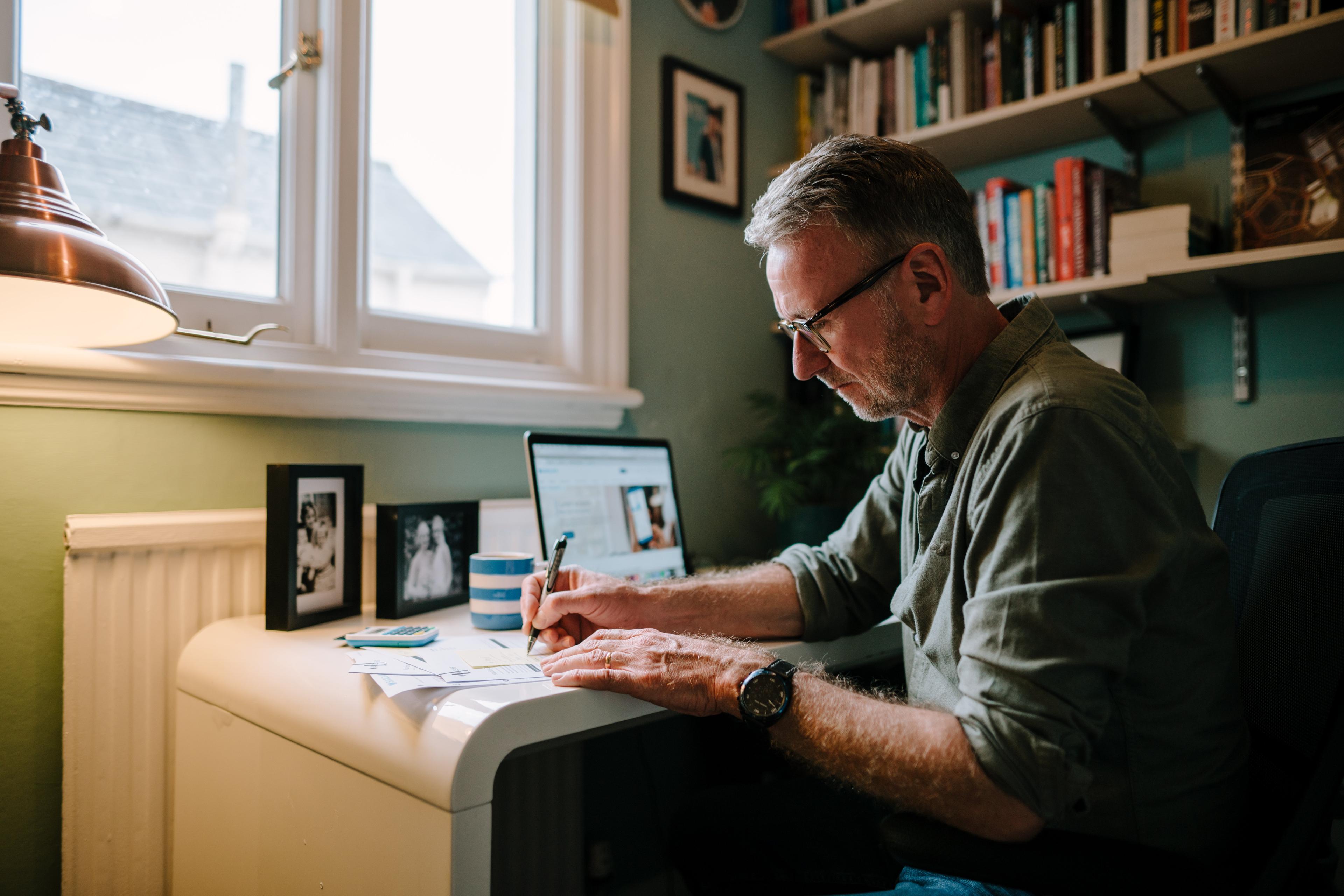 A middle-aged man is working at a desk in a cozy home office. He is writing on a document with an open laptop in front of him. The desk has framed photos, a mug, and a calculator. The background features a bookshelf filled with books. The scene conveys concentration and productivity.