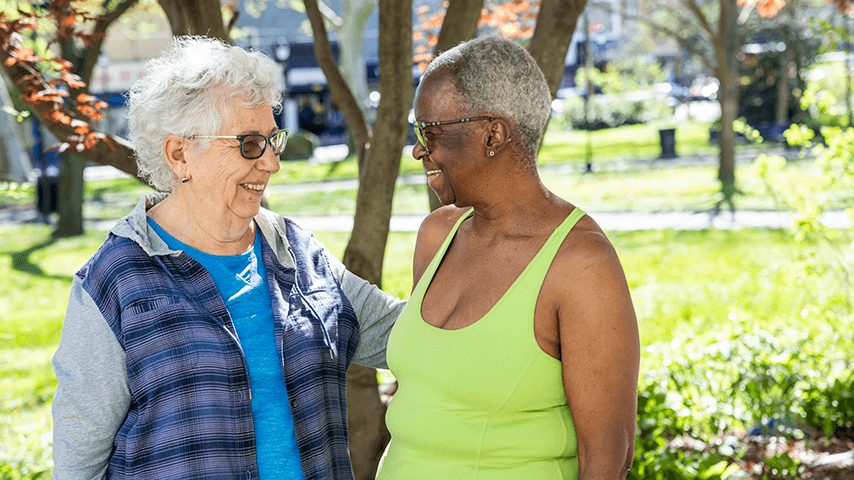 Two older women smiling and standing close together in a park. One is wearing glasses and a blue shirt, while the other is in a green tank top, with trees and greenery in the background.
