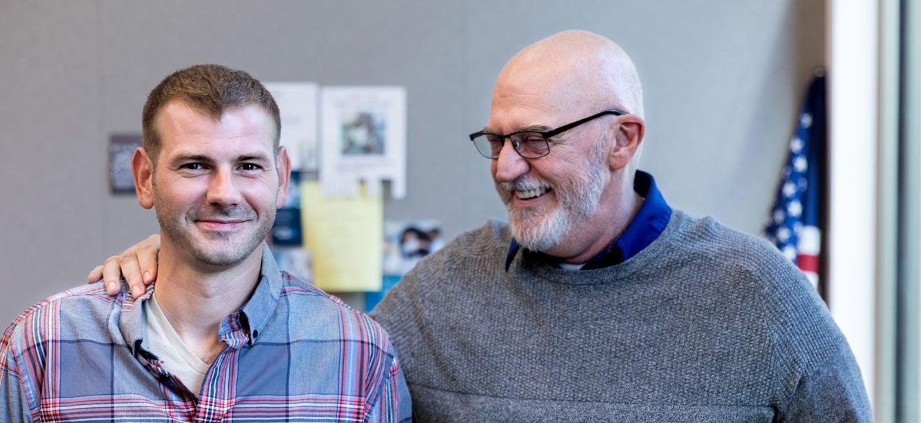 An older adult, senior father has his arm around his U.S. veteran son - both are smiling with pride.