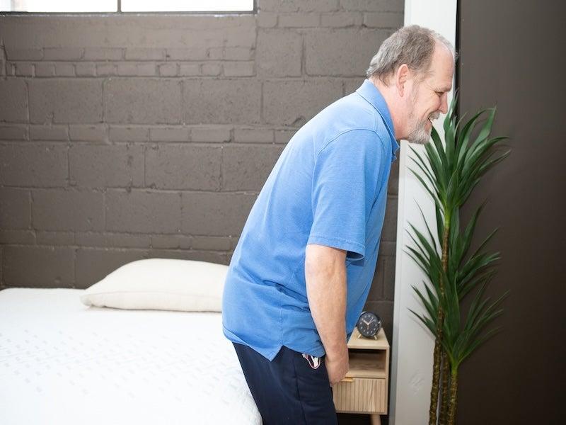 A man rises from sitting on the edge of the mattress.