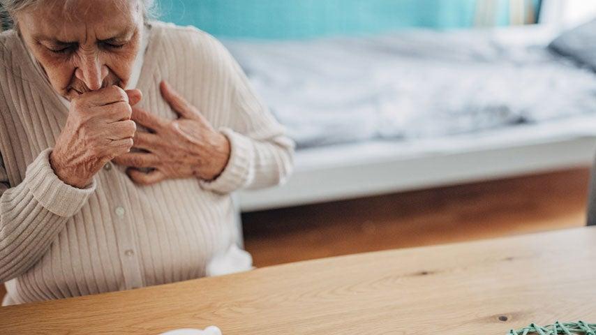 An older woman is coughing and touching her chest from her flu, while sitting at home.