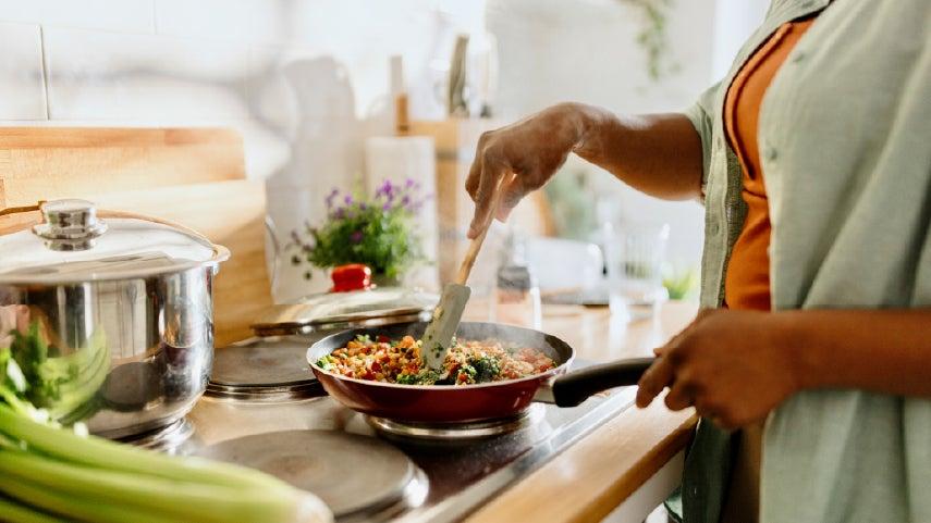 A black female caregiver is cooking stir fry on the stove, you can't see her face.