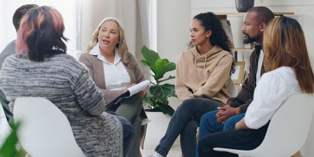 professionals sitting in chairs in a circle talking to a group leader holding a clipboard