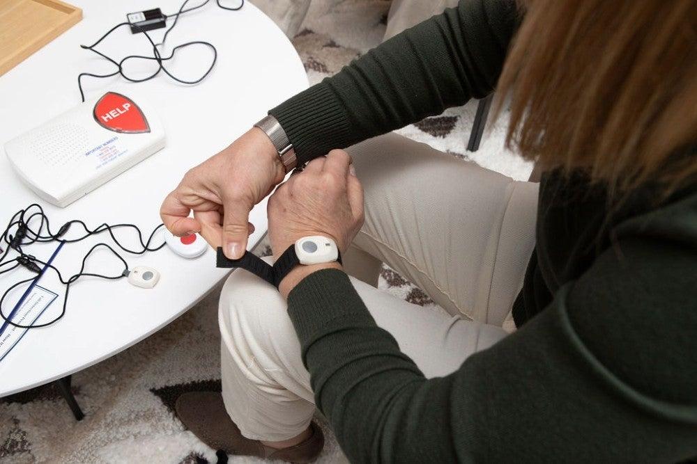 A woman sitting next to a medical alert system base station putting on a wristband with a help button