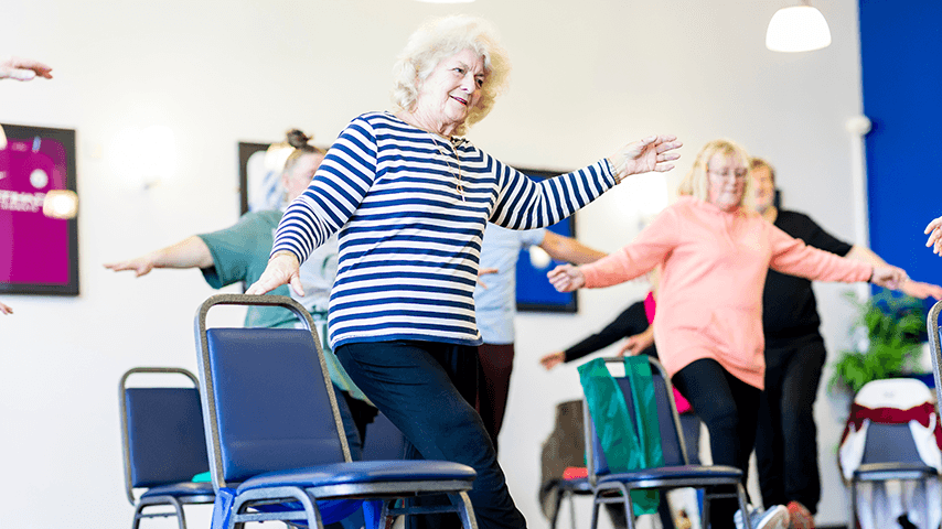 Older adults participate in a balance exercise class, using chairs for support during a standing exercise.
